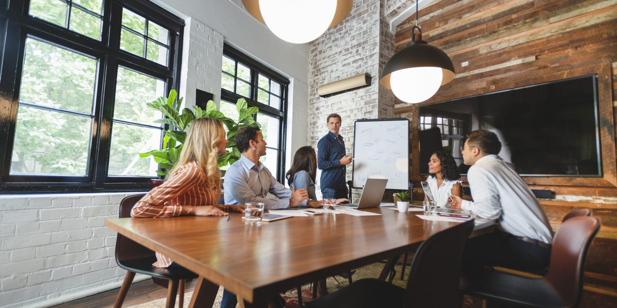 Business people watching a presentation on the whiteboard. A man is writing on the whiteboard. They are sitting in a board room, there are laptop computers and technology on the table. All are casually dressed. There is a brick wall behind him. Low angle view with copy space