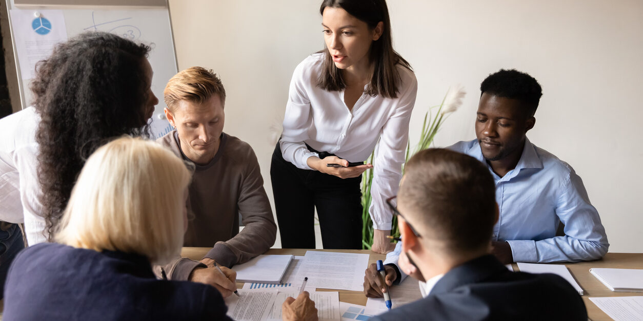 Female executive leader teaching diverse employees group at corporate office briefing analyze review paperwork marketing plan explain new strategy instructing workers mentoring at team office meeting