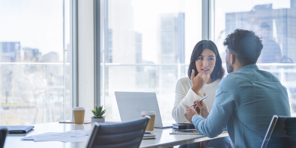 Business woman and man meeting and talking. Both are casually dressed. There is a laptop computer on the table. They are sitting at a boardroom table with a window behind them