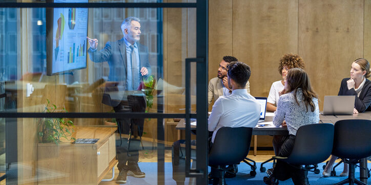 Mature man giving a big data presentation on a tv in a board room. There are several financial graphs and charts on the screen with a diverse group of people in the meeting room. There is paperwork and technology on the table