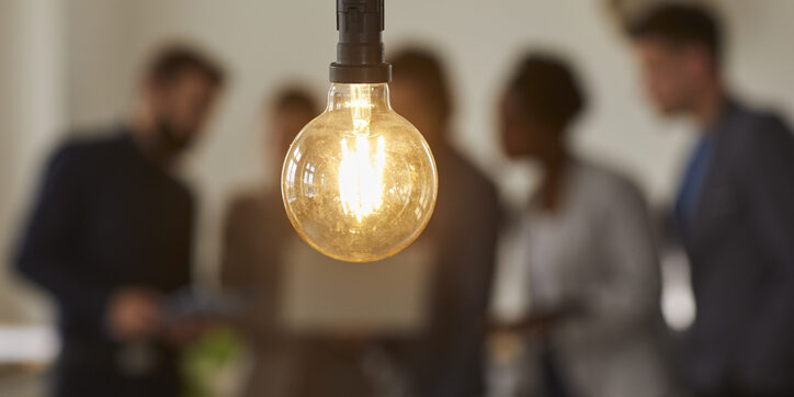 Glowing light bulb and business people over blurred background. Hanging light bulb on foreground, team of business people discussing ideas and brainstorming. Bright idea and creativity concept
